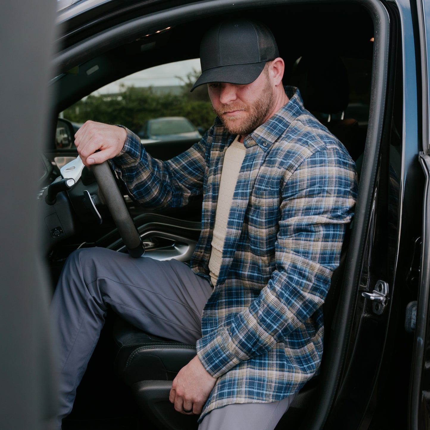 Man in a plaid shirt and cap sits in a car, gripping the steering wheel while wearing the RTS Tactical Body Armor Compression Shirt underneath. He looks focused, with the car door open, ready for action.